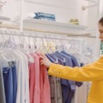 Woman browsing clothes on a rack in a clothing store, wearing a stylish yellow blazer.